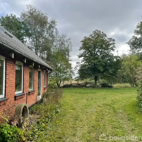 En rød murstensbygning med vinduer står stille i en frodig have med grønne træer og en græsplæne under en overskyet himmel.
