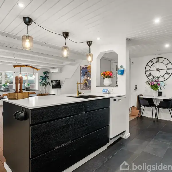 Køkken med sort-hvid køkkenø med tre hængende lamper, omgivet af spiseområde med sort ur på væggen. Blomster i vase og dekorationer skaber en hjemlig atmosfære.