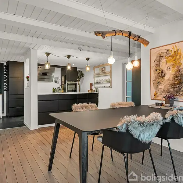 Sort spisebord med fire polstrede stole står i en spiseplads, omgivet af hvidmalede trælofter og kunstværk på væggen. Moderne loftslamper lyser rummet op ved siden af køkkenet.