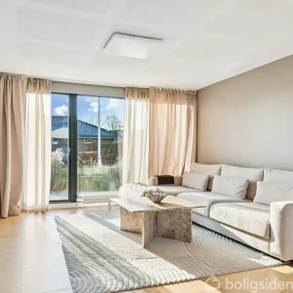 Lys stue med en stor, lysegrå sofa, placeret langs væggen. Sofabord i midten af en lys tæppe. Store vinduer med gardiner slipper dagslys ind og udsigt til en have.