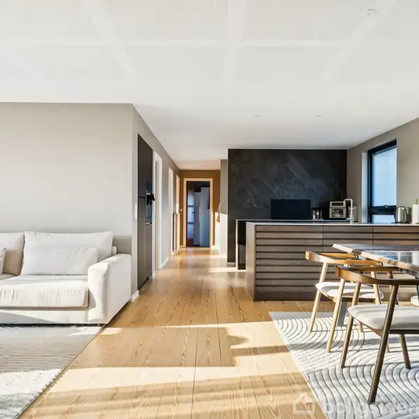 Stue med hvid sofa og sofabord til venstre, træbord og stole i midten, køkken med mørk bagvæg til højre. Sollys strømmer ind fra vinduet.