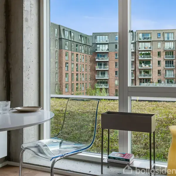 Et rundt hvidt bord står i et lyst rum ved siden af en gennemsigtig stol. Udenfor ses en række moderne bygninger. Bordet rummer en vase og nogle bøger.