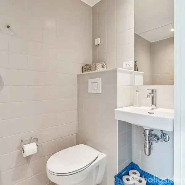 Toilet og håndvask i et moderne badeværelse. Toilettet er til venstre med toiletpapirholder monteret på væggen. Håndvasken er til højre under et spejl. Neutral flisebelægning.