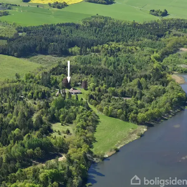 En bygning er omgivet af tæt skov og grønne marker. En flod løber gennem landskabet, som er frodigt og mangfoldigt, med en pil der peger på bygningen.