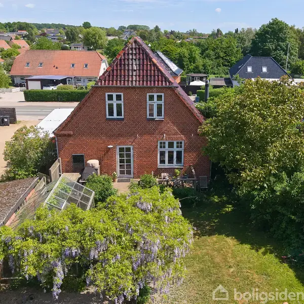 Murstenshus med rødt tag står i en have med træer og en blomstrende pergola. Huset har hvide vinduer, og er omgivet af grønne buske og nærliggende bygninger i et beboelsesområde.