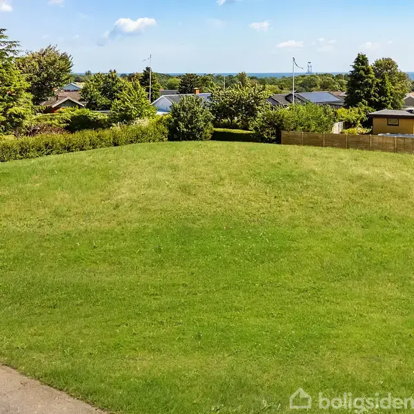 Grøn græsplæne ligger stille i et boligområde omgivet af huse og buske, med en enkelt lygtepæl stående på den ene side.