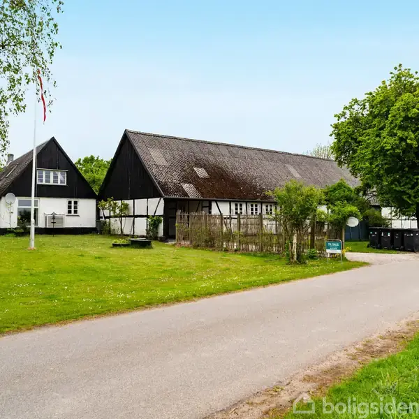 Hvid bindingsværksejendom med sort tag ligger ved en smal grusvej. Ejendommen er omgivet af grønne træer og omfatter også en flagstang med et hevet flag.