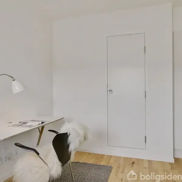 Et moderne skrivebord med en lampe og et magasin er placeret ved siden af en stol med fåreskind. Baggrunden er en hvid væg med en dør.