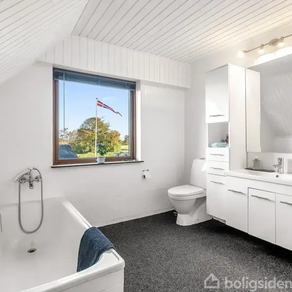 Badeværelse med badekar står ved siden af et toilet. Håndvask med skab på højre side. Et vindue viser en blå himmel og et dannebrogsflag udenfor.
