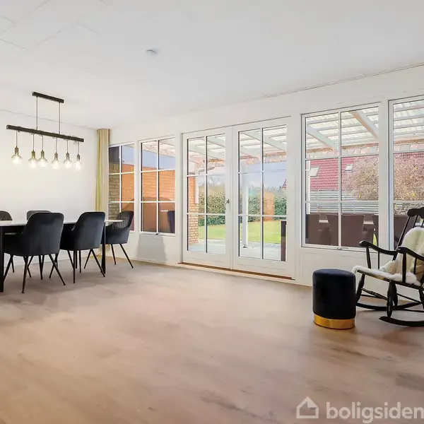 En spisestue med et sort spisebord og seks stole, står ved et stort vinduesparti, der fører ud til en have. Et gyngestol og en skammel står ved siden af.