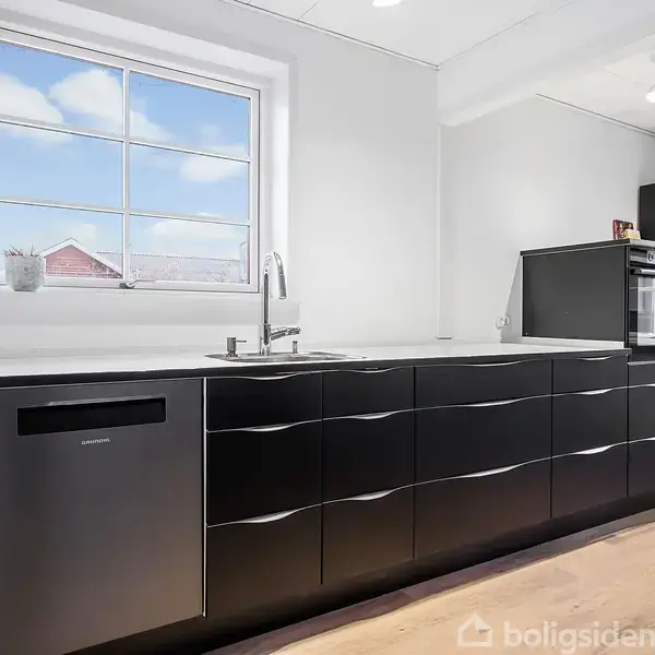 Sort køkkenbord står langs en væg, med indbygget vask og opvaskemaskine. Der er et vindue ovenover, som kaster lys ind i det modernt indrettede rum.