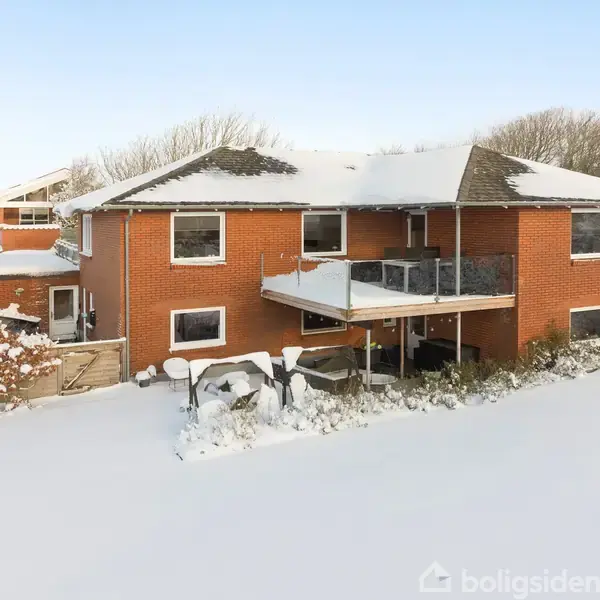 Rødt murstenshus med snebeklædt tag står stille i en snefyldt have. En hævet terrasse med glasrækværk er tilknyttet huset. Omgivet af sneklædte buske og træer.