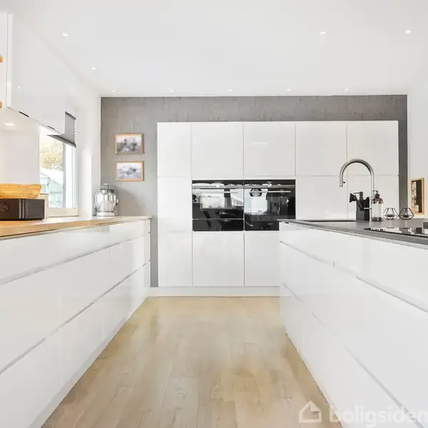 Et køkken med hvide skabe og træbordplader, indeholder kaffemaskine og diverse køkkenredskaber. Rummet er lyst og moderne med en centralt placeret vask og ovn indbygget i skabene.