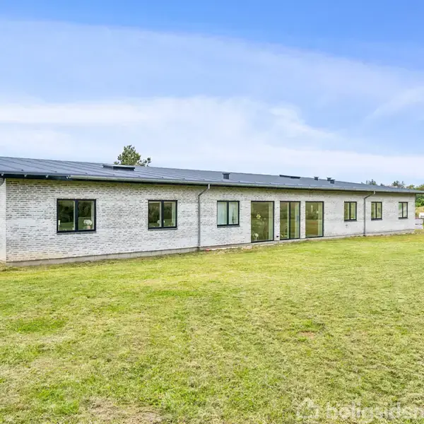 En langstrakt, moderne en-etagers bygning med fladt tag står stille på en stor græsplæne. Vinduerne er jævnt fordelt langs facaden. I baggrunden ses en åben, grøn landskab.