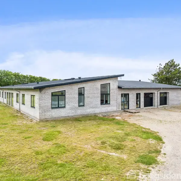 En moderne, enkeltliggende bygning med fladt tag, omgivet af åbent græsareal og træer i baggrunden under en klar blå himmel.