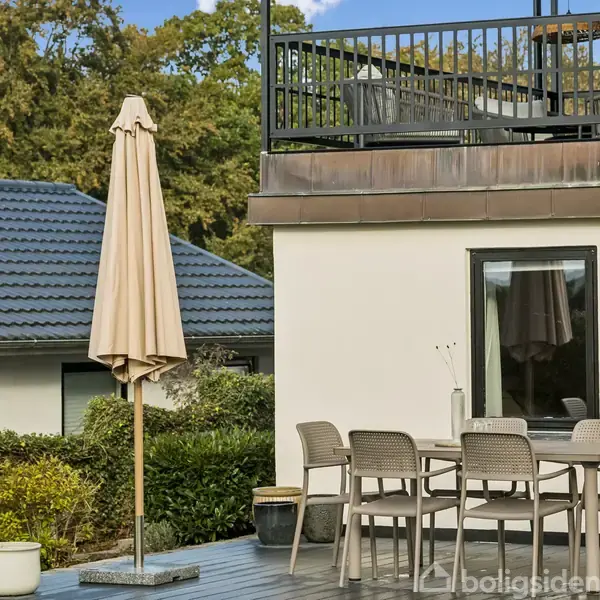 En terrasse med et bord og seks stole står ved siden af en hvid bygning. En stor parasol er rejst, og der er grønne buske og et hus med mørkt tag i baggrunden.