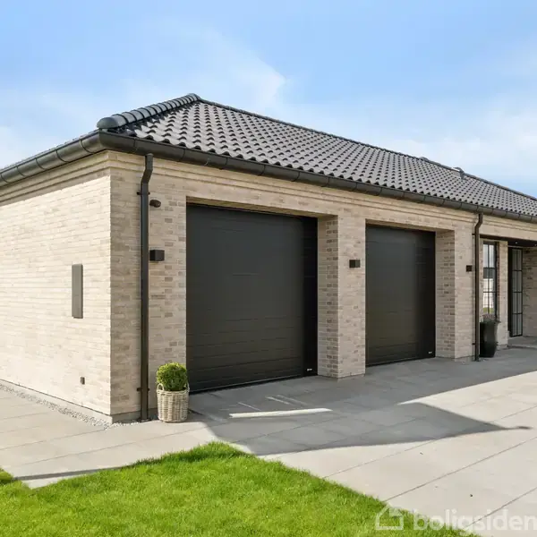 Et moderne enfamiliehus med fladt tag, to garageporte og lyse mursten, står på en velplejet græsplæne med betonfliser i solen, omgivet af åbne, grønne marker.