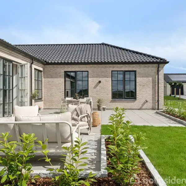 En moderne terrasse med lyse havemøbler står foran et murstenshus med sort tegltag. Grønne planter og en velplejet græsplæne omgiver området, under en blå himmel.