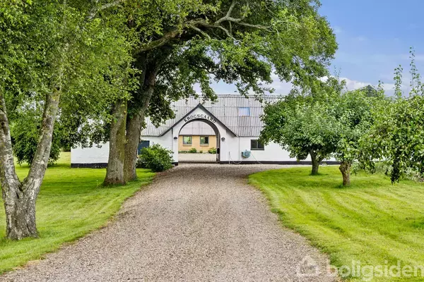 Hvid bondegård med en grusvej foran, omgivet af træer og græs. Tekst på gårdens gavl: "TROSCAMIN".