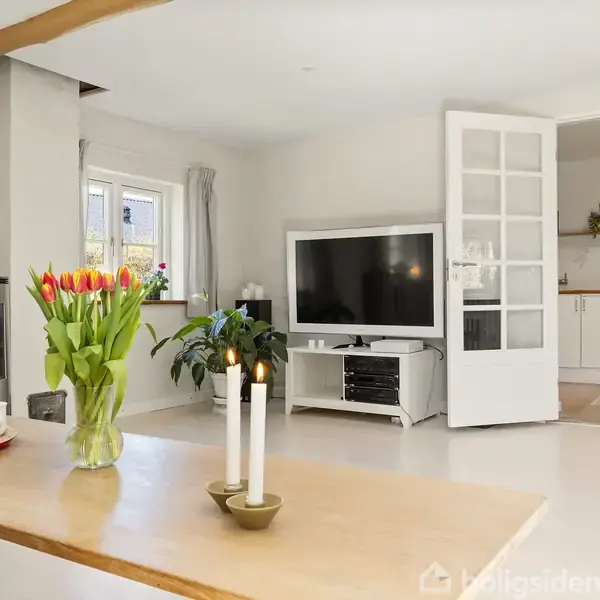 Et moderne stueinteriør med et stort TV på en stand. Et spisebord med blomster og stearinlys i forgrunden. En brændeovn i hjørnet ved et vindue, danner en hyggelig stemning.