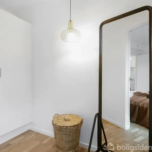 Et spejl står oprejst i et lyst rum med trægulv. En kurv er placeret ved siden af spejlet. En lampe hænger ned fra loftet, og et skab er i baggrunden.