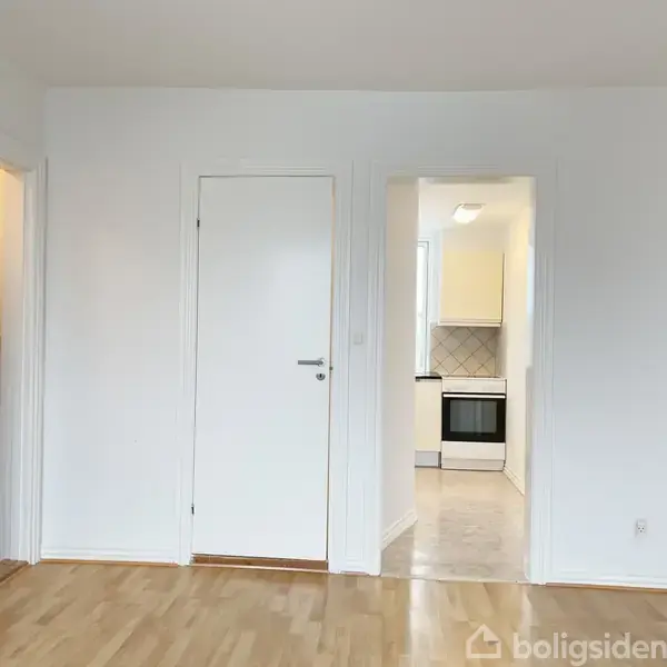 En hvid dør er lukket mod en hvid væg, ved siden af en åbning ind til et køkken med komfur. Trægulv dækker rummet, og der er en person til venstre.