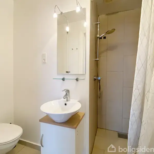 En moderne badeværelsesindretning med en rund hvid håndvask på et træskab, en vægmonteret spejl over vasken, et toilet til venstre og en bruser i baggrunden.