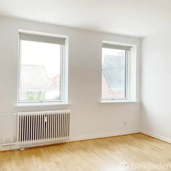 To vinduer med rullepersienner, står uåbnede, og lyser op et tomt rum med lyse trægulve, mens en radiator sidder under det venstre vindue.
