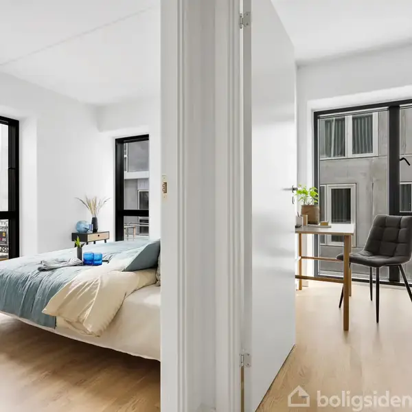 Soveværelse med seng og blå sengetæppe, tilstødende arbejdsværelse med skrivebord og stol. Lys træparketgulv, store vinduer med udsigt til en bygning med flere etager.