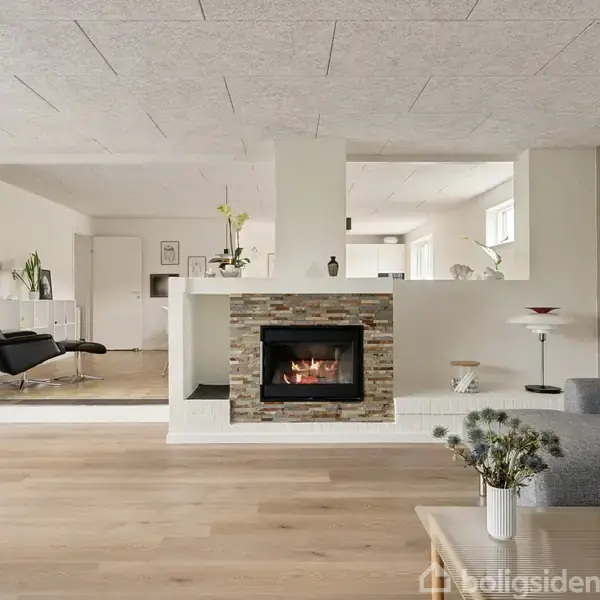 En moderne stue med en central pejs, flammer synlige, omkranset af lys indretning. Sofa og planter tilføjer komfort. Rummets åbne plan skaber en sammenhængende og lys atmosfære.
