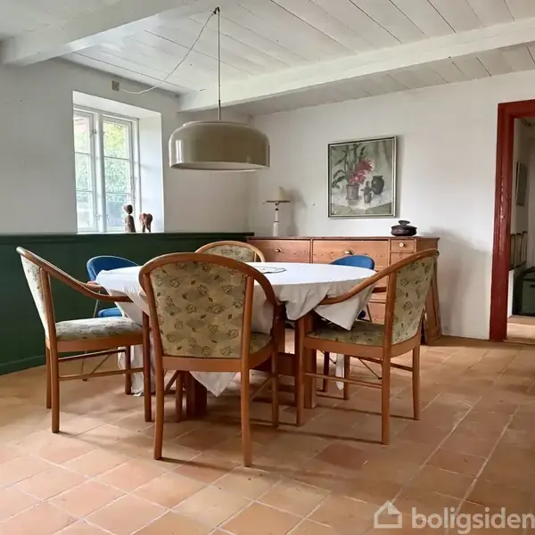 Et rundt bord med hvid dug er omgivet af seks polstrede stole i en spisestue. Baggrunden har en kommode, en lampe, et maleri og grønne paneler.