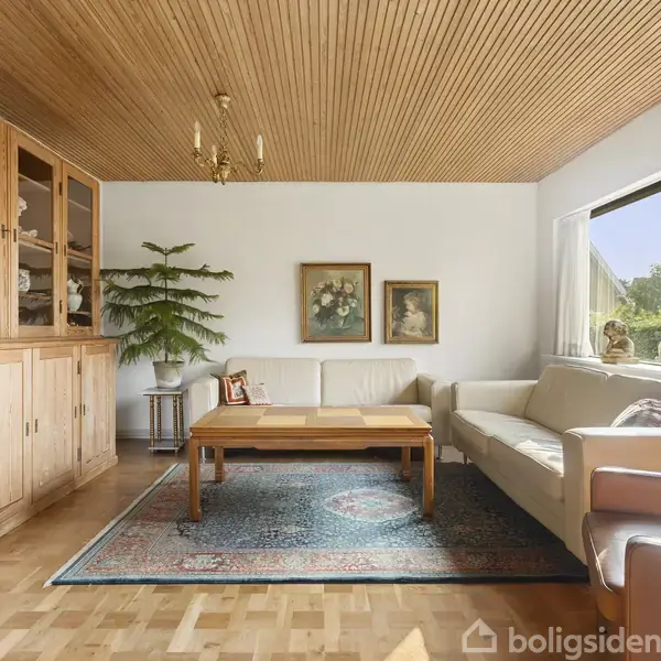 Et lyst og rummeligt stueområde med lyse lædersofaer og et sofabord, omgivet af en tæppebelagt parketgulv. Et træskab og grønne planter dekorerer rummet, der har billeder på væggen.