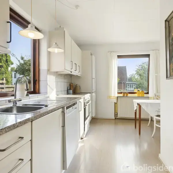 Et lyst køkken med hvide skabe og en grå granitbordplade. To vinduer giver udsigt til en grøn have. Et spisebord står ved vinduet, og et farverigt maleri hænger på væggen.