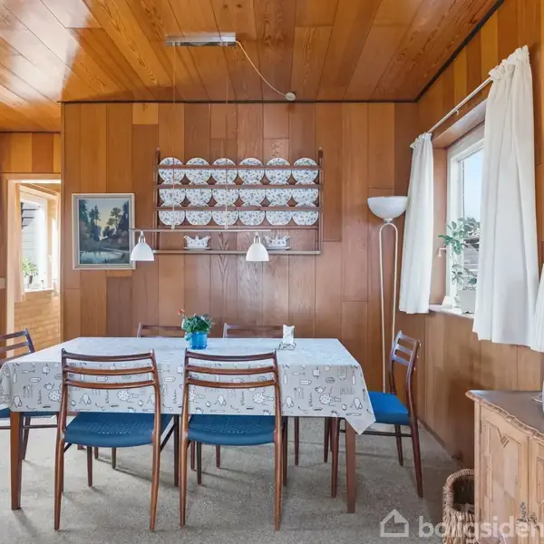Spisebord med fem stole står i et rum med træpaneler på vægge og loft. To lamper hænger over bordet. En hylde med porcelæn pynter væggen. Vindue med gardiner til højre.
