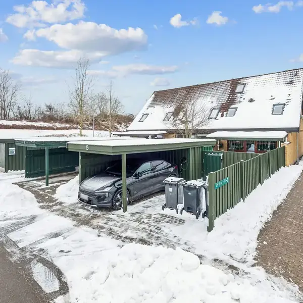 En bil holder parkeret under en carport ved et rækkehus med snedækket tag. Omkring bil og hus er der hegn og snebeklædte stier. Skraldespande står tæt ved carporten.