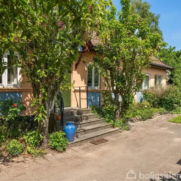 Et hus med beige facader står omgivet af frodige træer og buske. En trappe ved indgangen fører til en blå vase. En græsplæne ligger til højre.