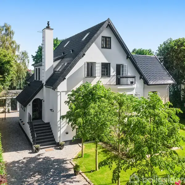 Et hvidt, to-etagers hus med sort tag står omgivet af grønne træer og en velplejet have. Huset har en balkon, skodder på vinduerne og en bred trappe ved indgangen.