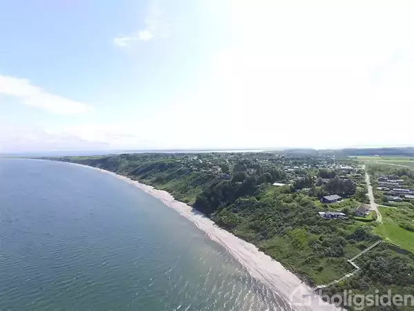 Kystlinjen omfatter en sandstrand skrånende mod det rolige hav, mens grønne bakker og spredte huse ligger bagved. Landskabet strækker sig mod horisonten under en klar, blå himmel.