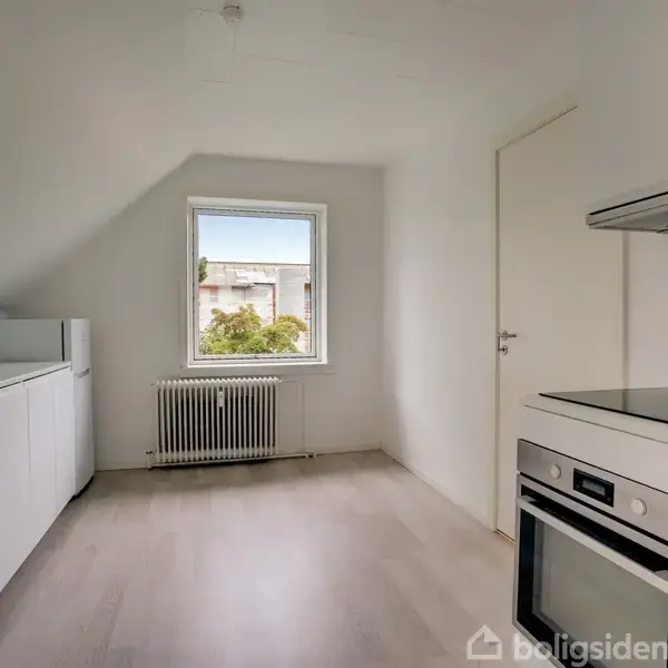 Et moderne køkken med hvide skabe står langs væggen. En indbygget ovn og kogeplade er til højre. Et vindue i midten giver udsigt til træer udenfor.