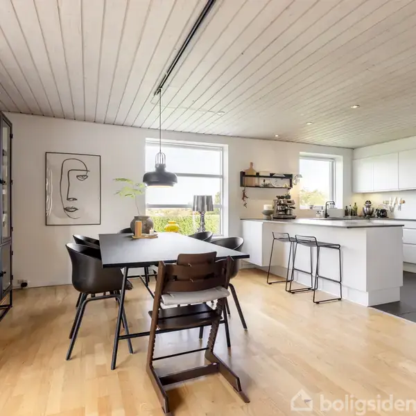 Et spisebord med stole står i et moderne køkken-alrum. En dekorativ vaseskulptur og kunst på væggen skaber en stilfuld atmosfære. Køkkenet er lyst med hvide skabe.
