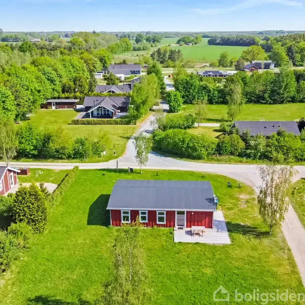 Rødt hus med fladt tag står på grøn græsplæne i et landområde. Omkring huset er der træer og små veje. Andre huse i afstand med skov og marker i baggrunden.