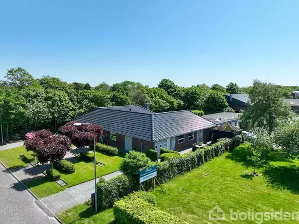 Et parcelhus med gråt tag står omgivet af en velholdt have med træer og buske. Huset ligger ved siden af en vej, og der er skov i baggrunden.