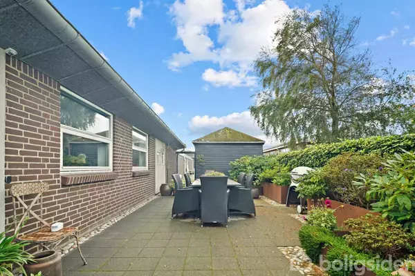 En terrasse med et bord og sorte stole står ved siden af en murstensbygning. Haven er frodig med buske og et stort træ. Himlen er blå med skyer.