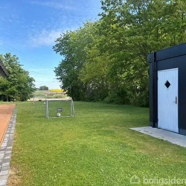 En græsplæne med et lille fodboldmål står mellem to bygninger. Omkring er der træer og en blå himmel i baggrunden.