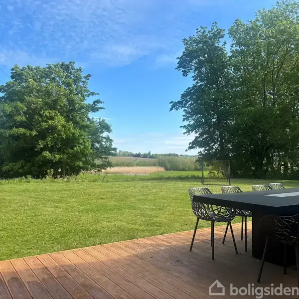 Et sort havebord med stole står på en træterrasse. I baggrunden er en græsplæne omkranset af store træer under en blå himmel.