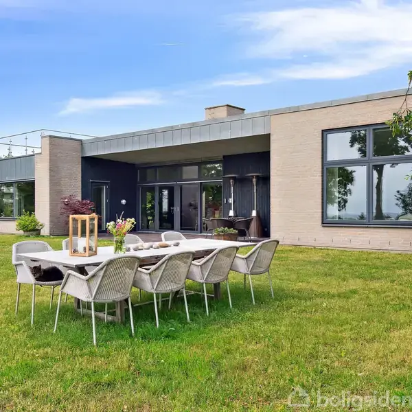Et moderne hus står på en velholdt græsplæne. Foran huset er et bord arrangement med stole, nogle pyntet med lanterner og blomster.