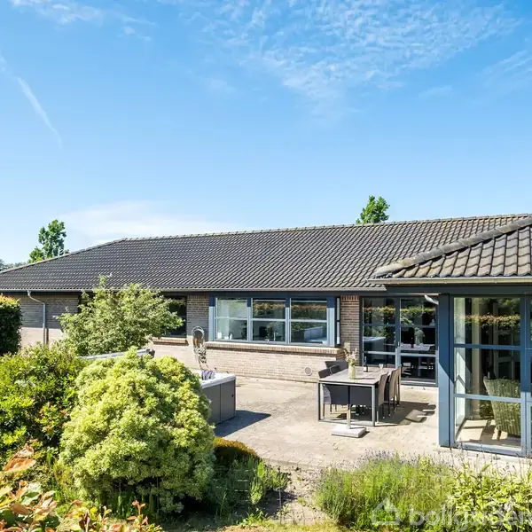 Et murstenshus med sorte tagsten står i en solrig have. Udendørs terrasse møbleret med et bord og stole. Grønt buskads omgiver terrassen under en klar blå himmel.