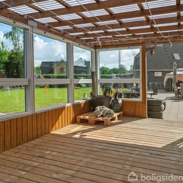 Overdækket terrasse med træbjælker og glasvægge, der giver udsigt til en grøn have. Udendørs møbler og planter er placeret på flisebelagt område ved siden af plænen.