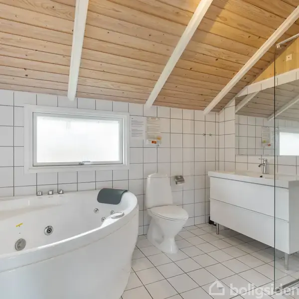 Boblebadkar står i et lyst badeværelse med hvidklinker på væggene og gulvet. Ved siden af er et toilet, håndvask med skab, spejl og brusekabine i et rum med træloft.