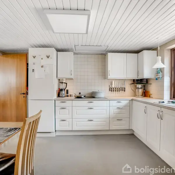 Hvidt køkken står stille. Skabene og skufferne er moderne, mens træbordet med stole sidder til venstre. Store vinduer viser en grøn udsigt udenfor.